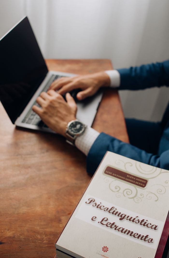 A professional in a suit types on a laptop next to a book in a well-lit setting.