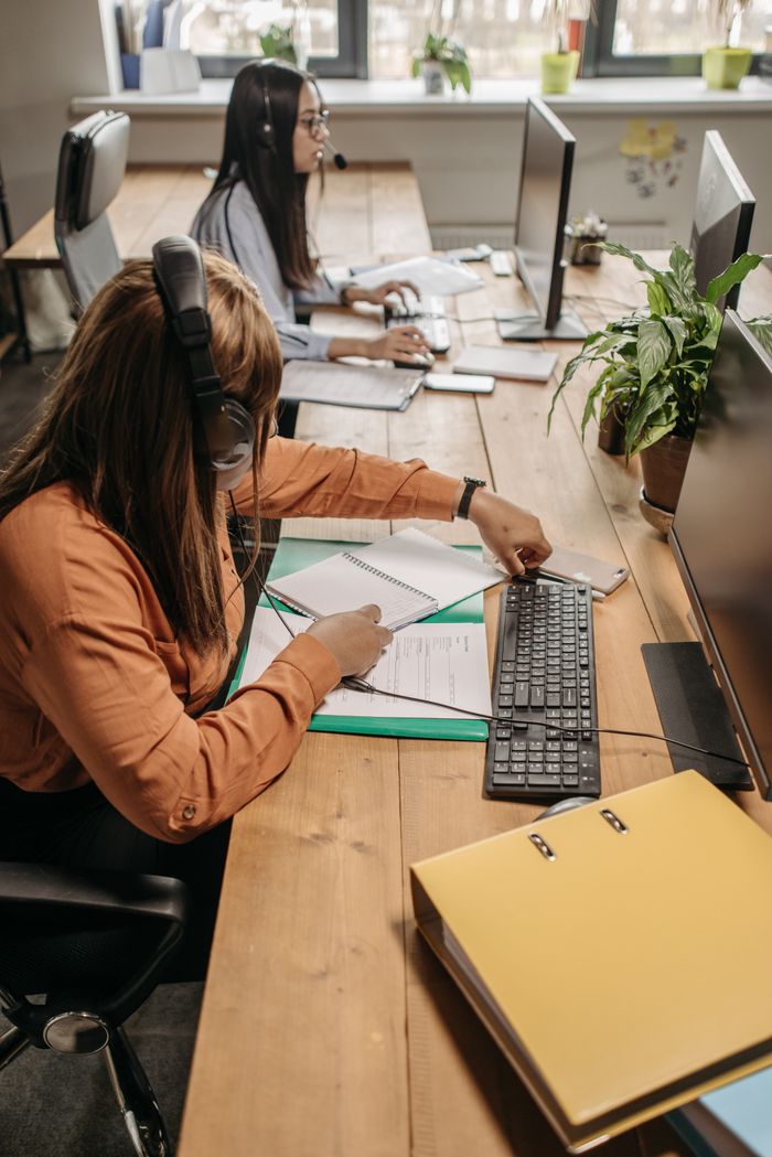 Two employees working at desks in a modern office, using computers and taking notes.